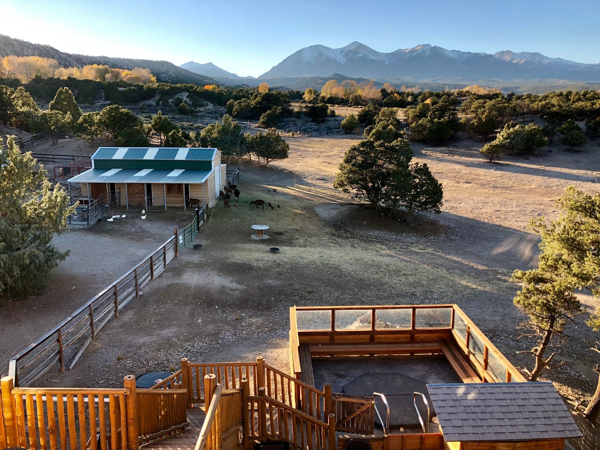 Wooden deck with hot tub overlooking a ranch and mountains on a sunny day.