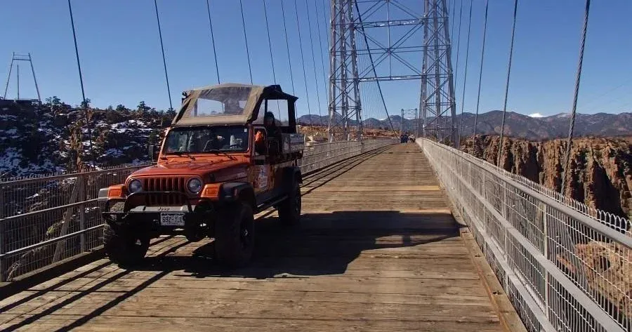 Orange Jeep driving across a suspension bridge with mountains in the background under a blue sky.