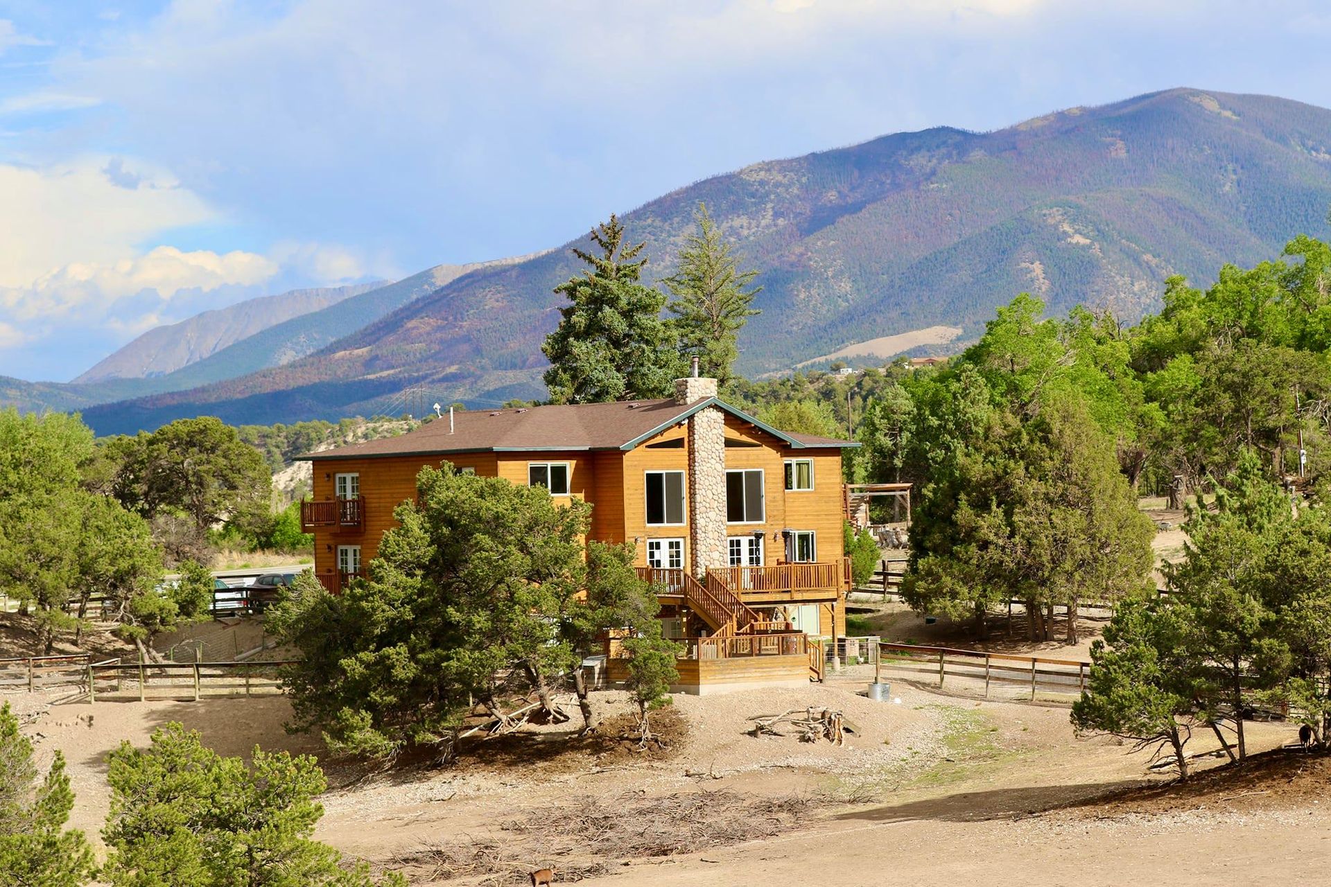 Two-story wooden house with deck, surrounded by trees, mountains in background.