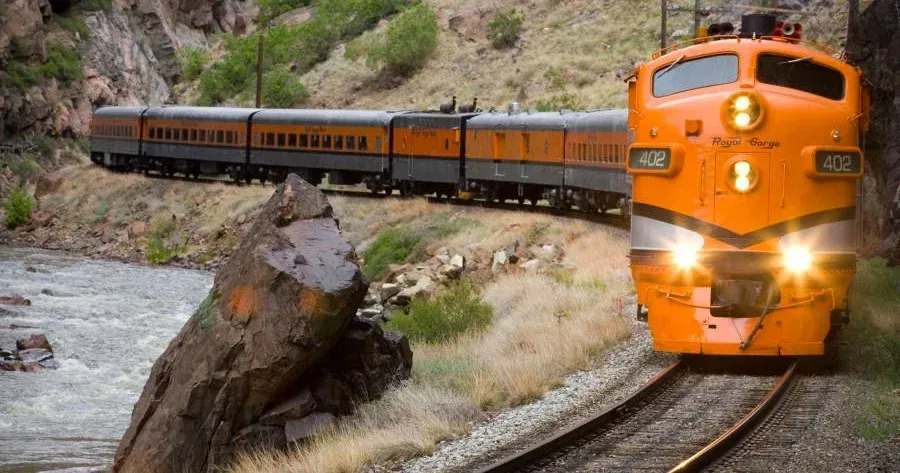 Orange train traveling on tracks alongside a river, passing a rocky landscape.