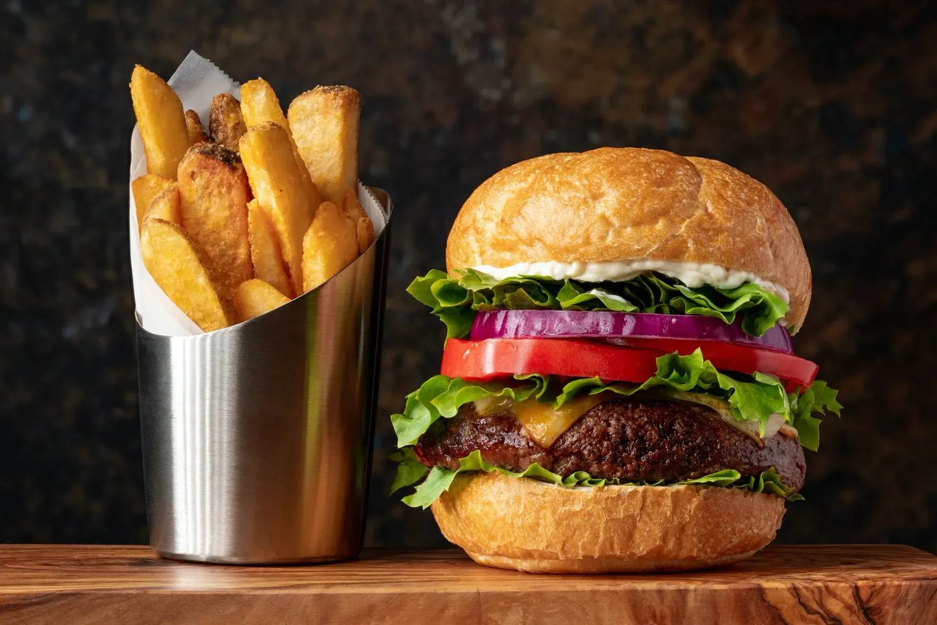 Burger with tomato, red onion, lettuce, and cheese, next to a metal cup of fries on a wooden board.
