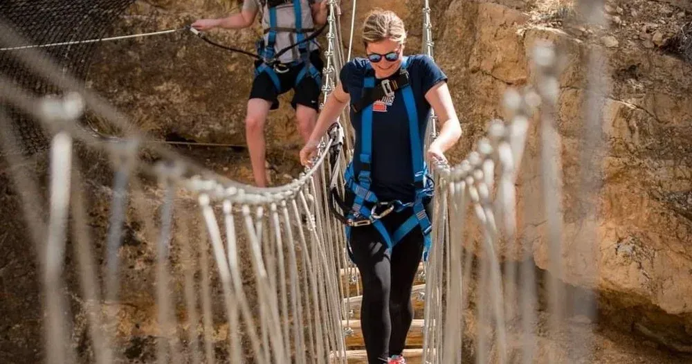 Woman in harness walks rope bridge; another person follows. Brown rock walls.