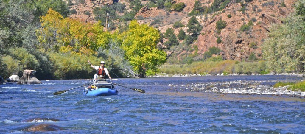A person in a raft on a blue river, rowing towards the viewer, surrounded by green and yellow trees and brown mountains.