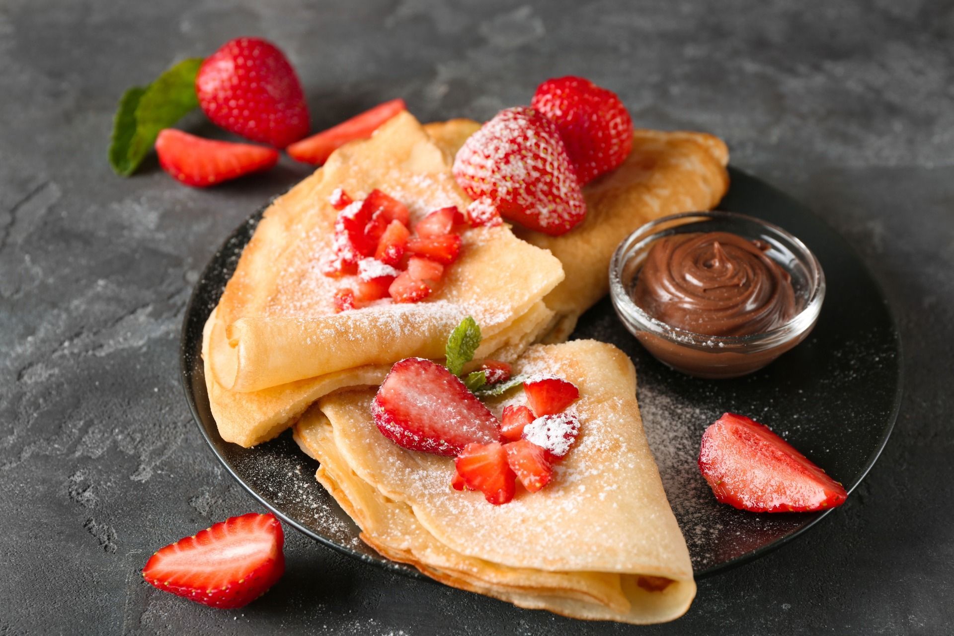 Crepes with strawberries and chocolate sauce on a black plate, sprinkled with powdered sugar.
