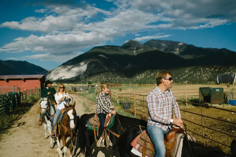 People riding horses on a dirt path, mountain backdrop, blue sky.