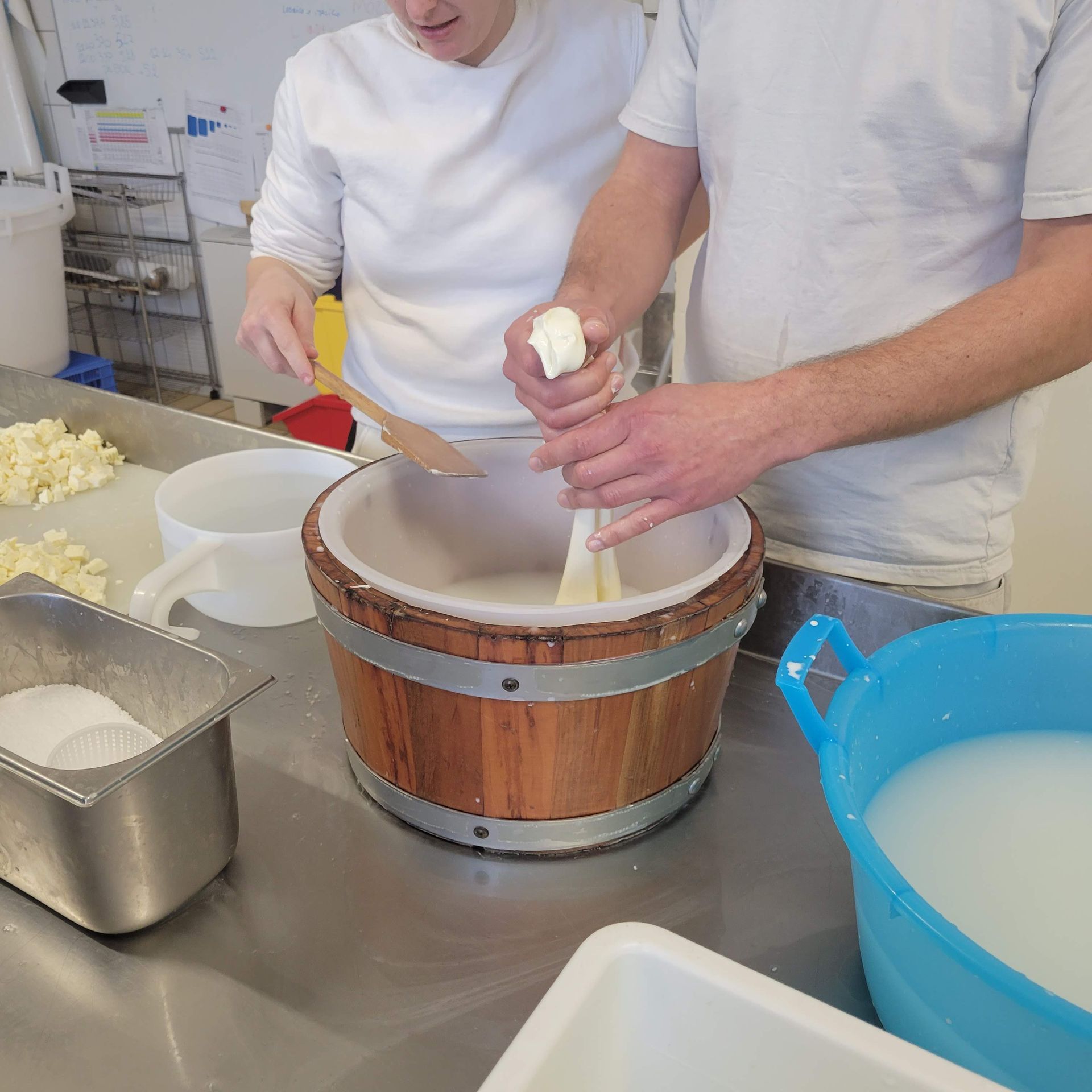 Two people making cheese in a kitchen, using wooden bucket and tools.