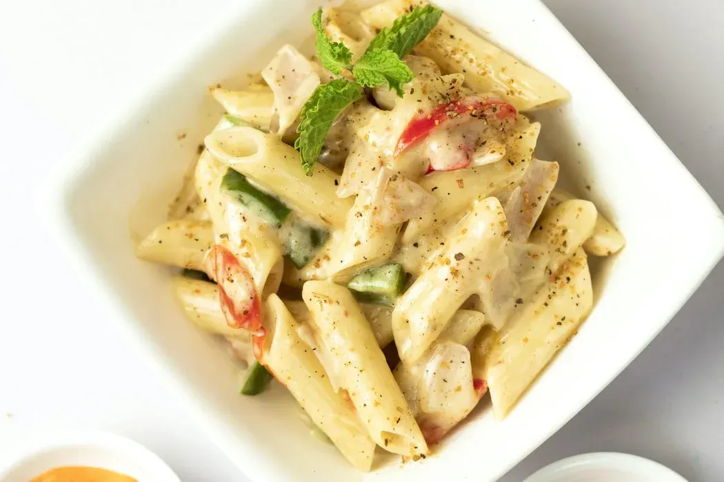 Penne pasta with a creamy sauce, red and green peppers, and a sprig of mint in a square white bowl.