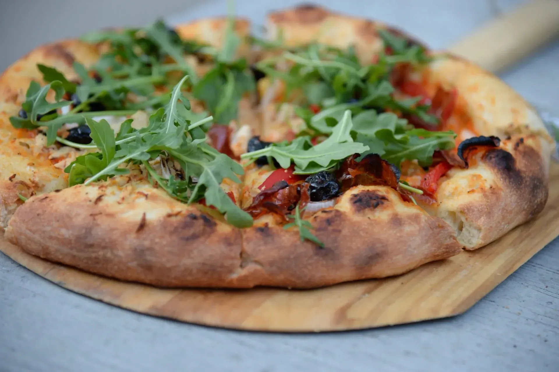 Pizza with arugula, roasted tomatoes, and olives on a wooden peel.