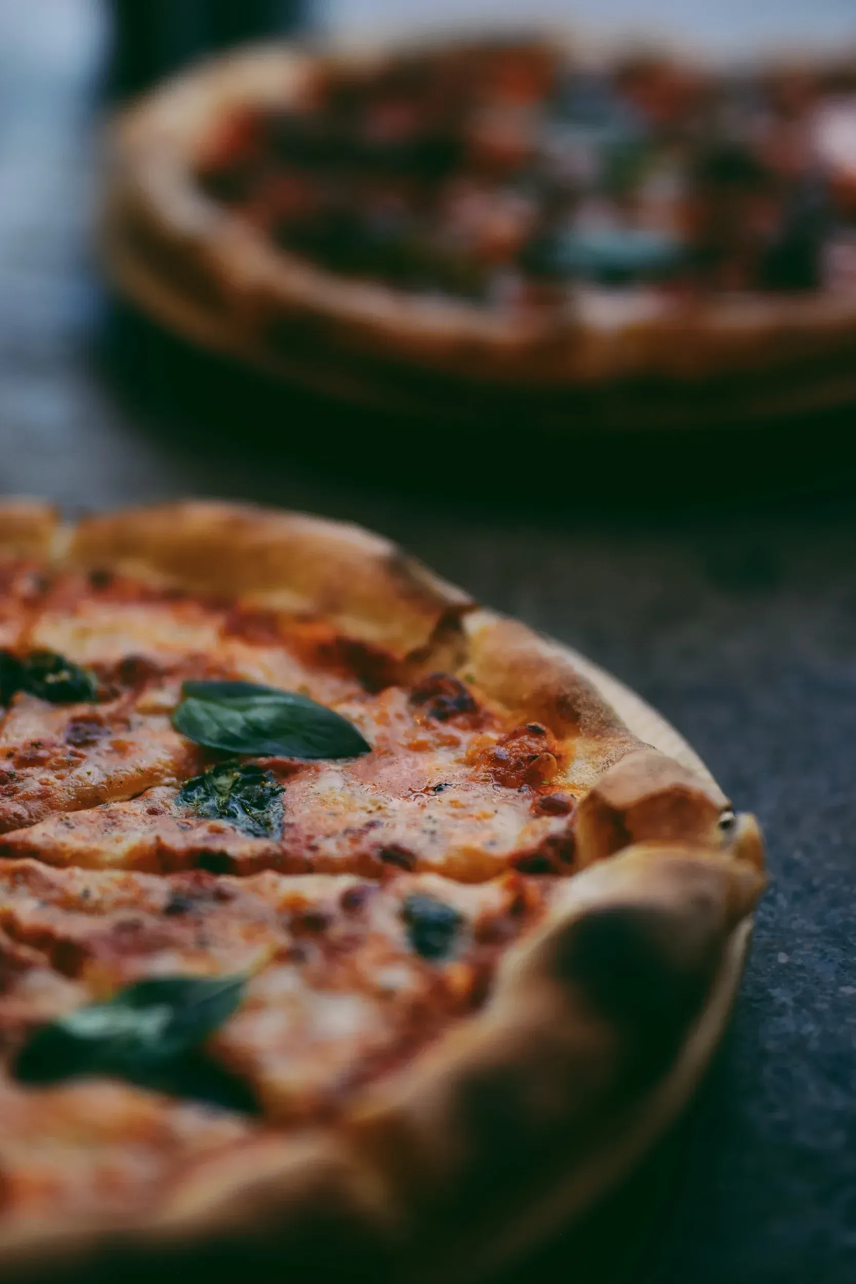 Two pizzas, one in focus. Crispy crust, toppings, basil leaves visible.