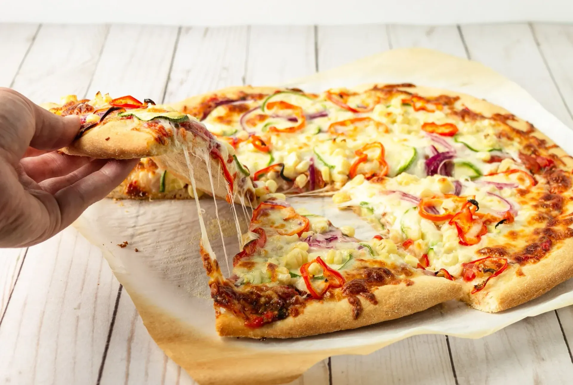 Hand lifting a slice of pizza with melted cheese, revealing toppings on a wooden surface.