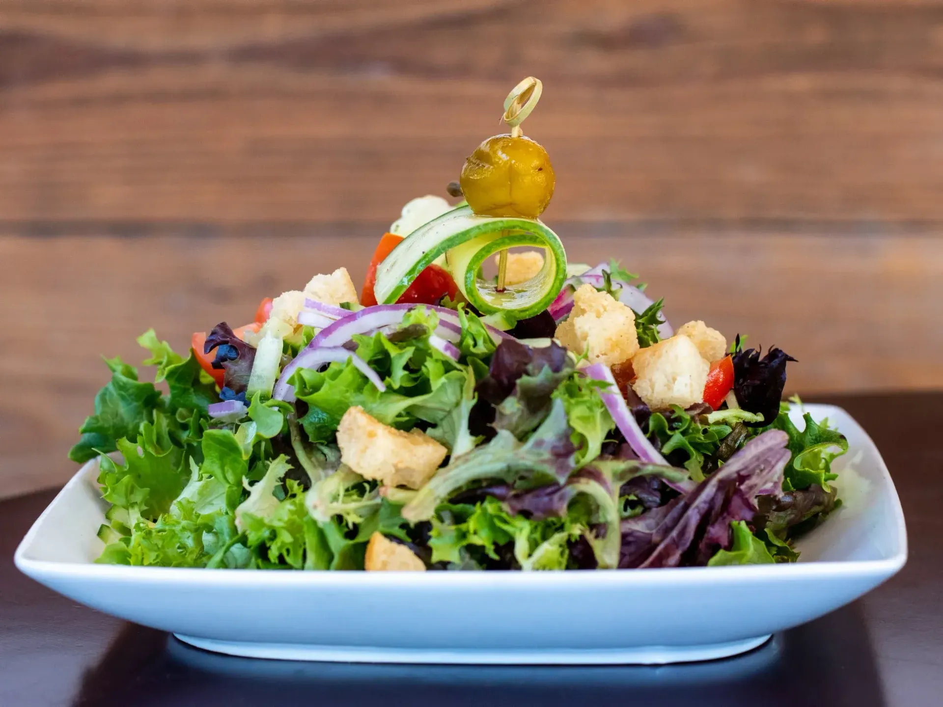 Salad with greens, croutons, red onion, cucumber, and cherry tomatoes, topped with a skewer and a pickle.