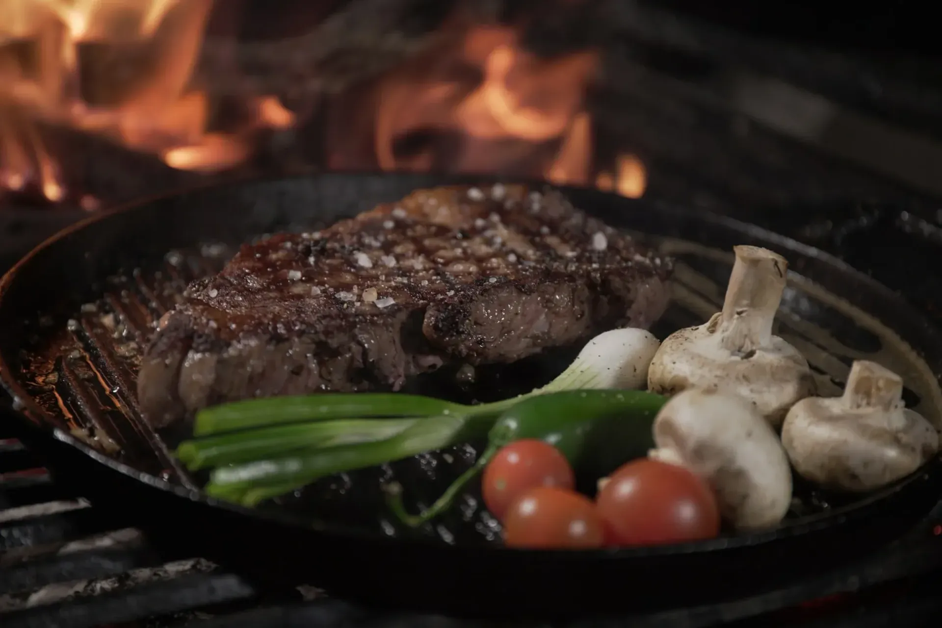 Steak, vegetables, and mushrooms in a pan on a grill with flames in the background.