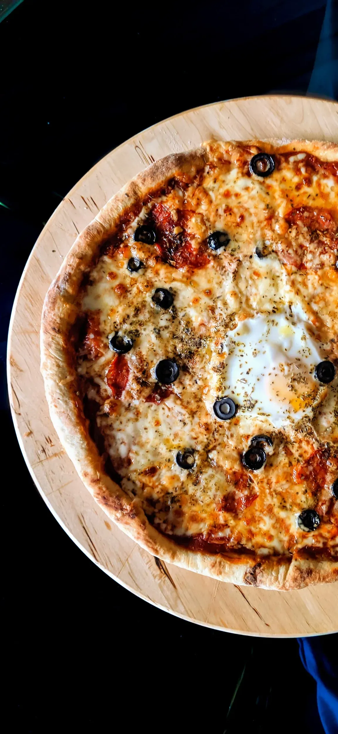 Pizza with cheese, olives, and an egg on a wooden board against a dark background.