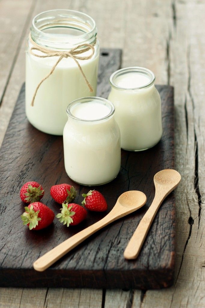 Three jars of yogurt, strawberries, and wooden spoons on a wooden cutting board.