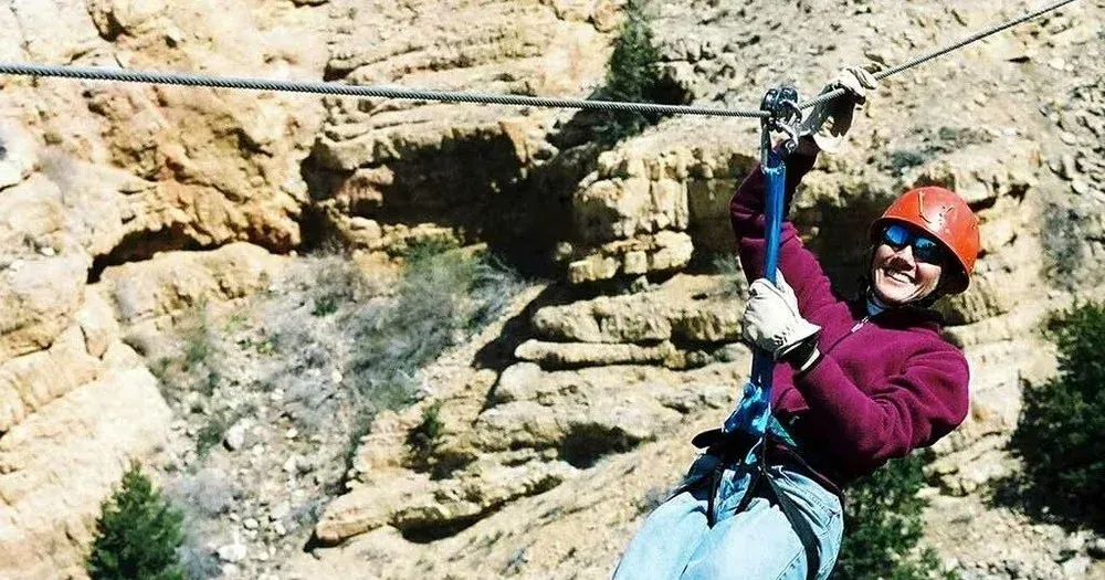 Person on a zip line, wearing a helmet and sunglasses, smiling against a rocky cliff backdrop.