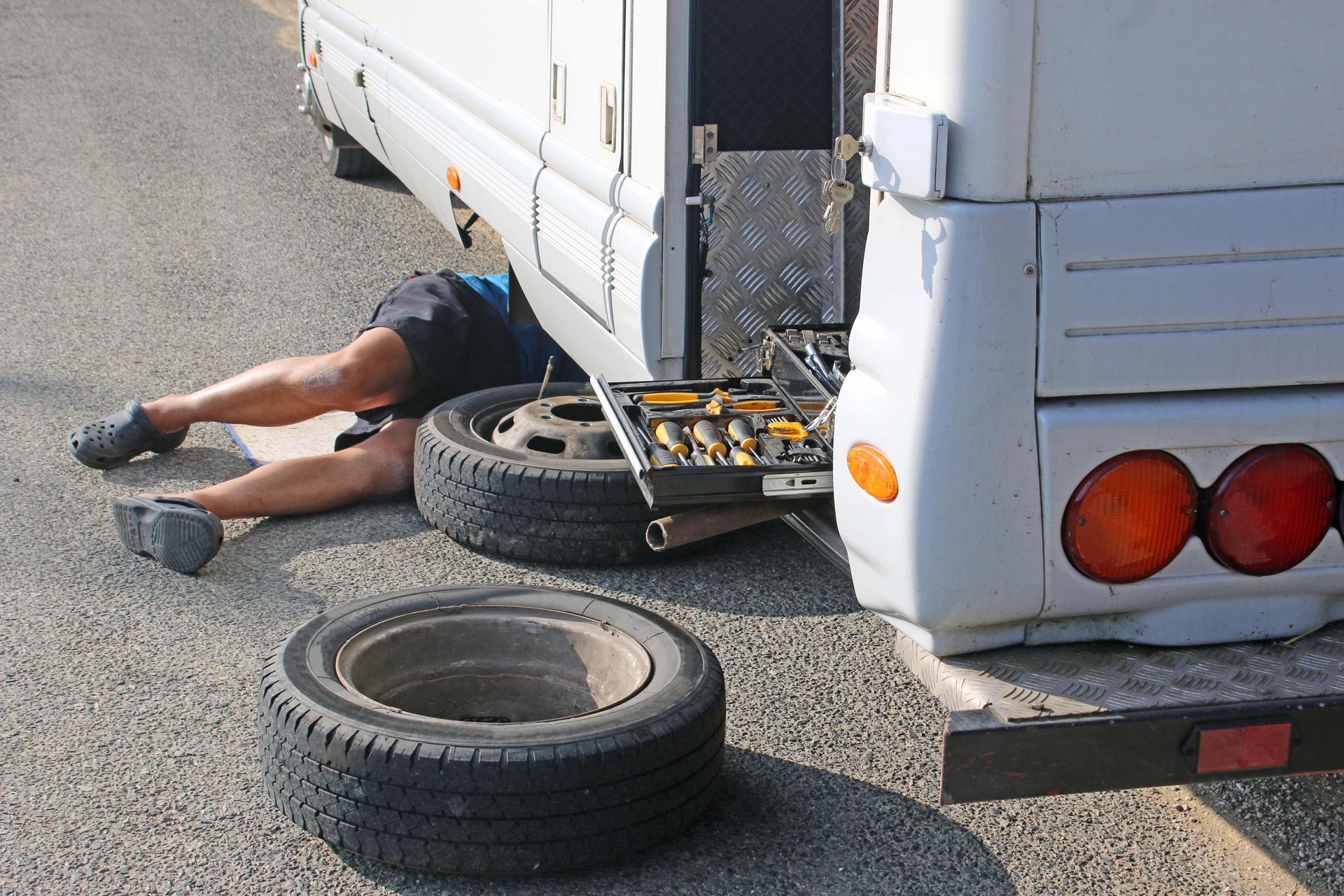 Technician performing mobile RV repair services under a motorhome beside a spare tire and tool kit.