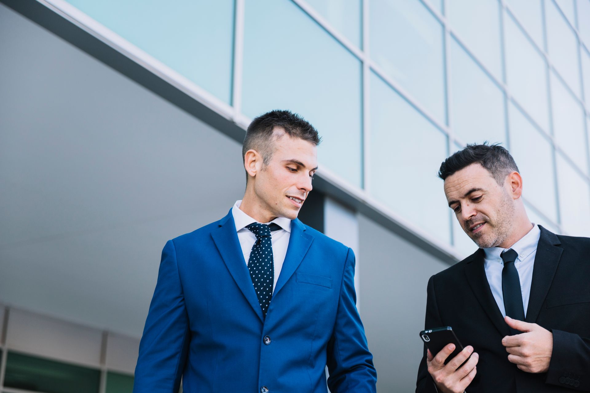 Two men in suits look at a phone in front of a modern building.
