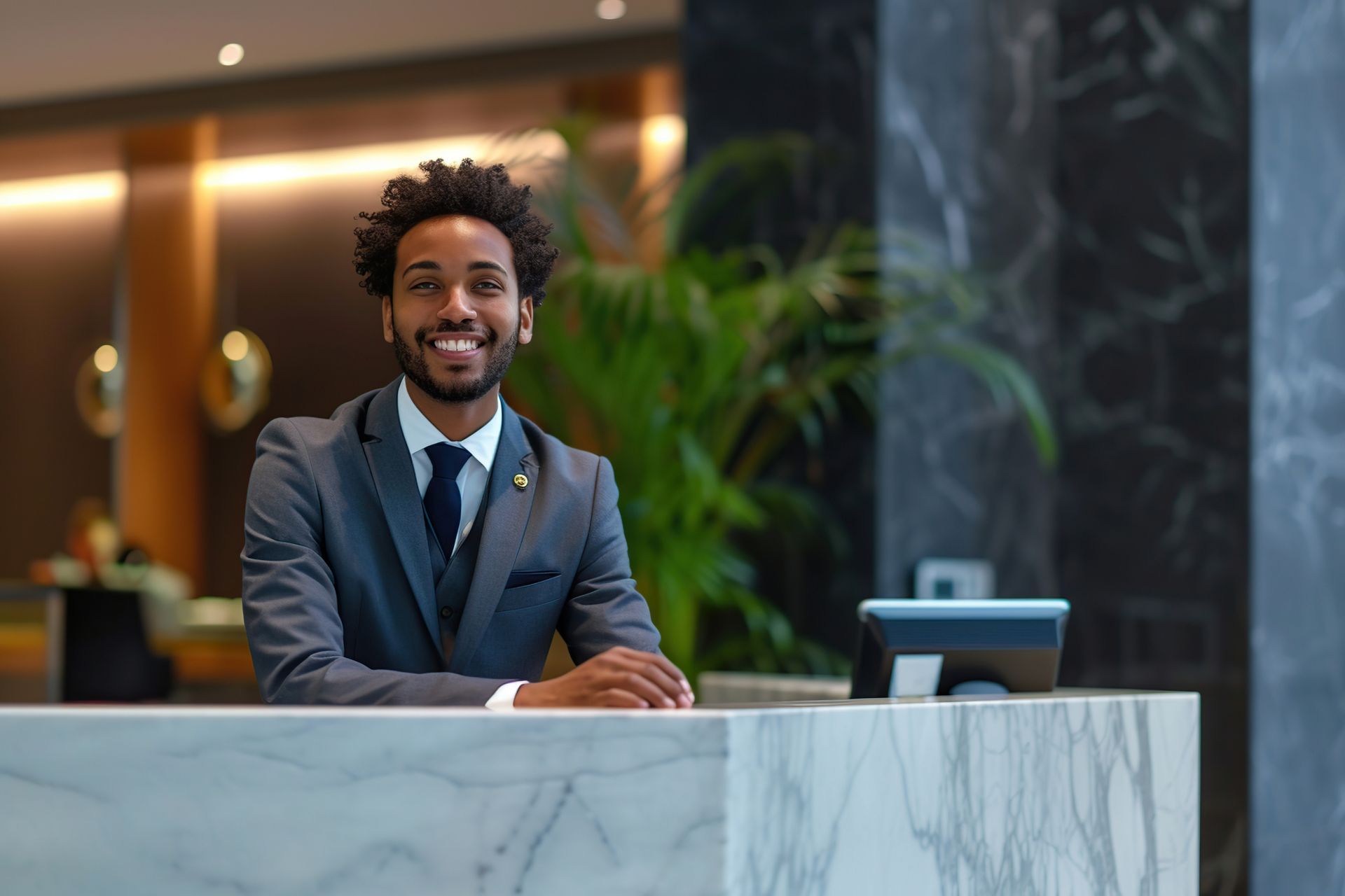 Smiling hotel receptionist at a marble desk in a lobby, wearing a suit and tie.