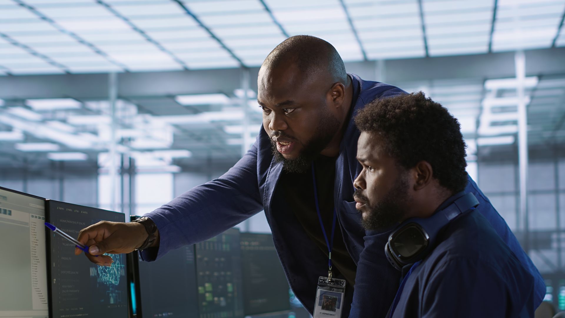 Two people in a server room looking at computer screens, one pointing with a pen.