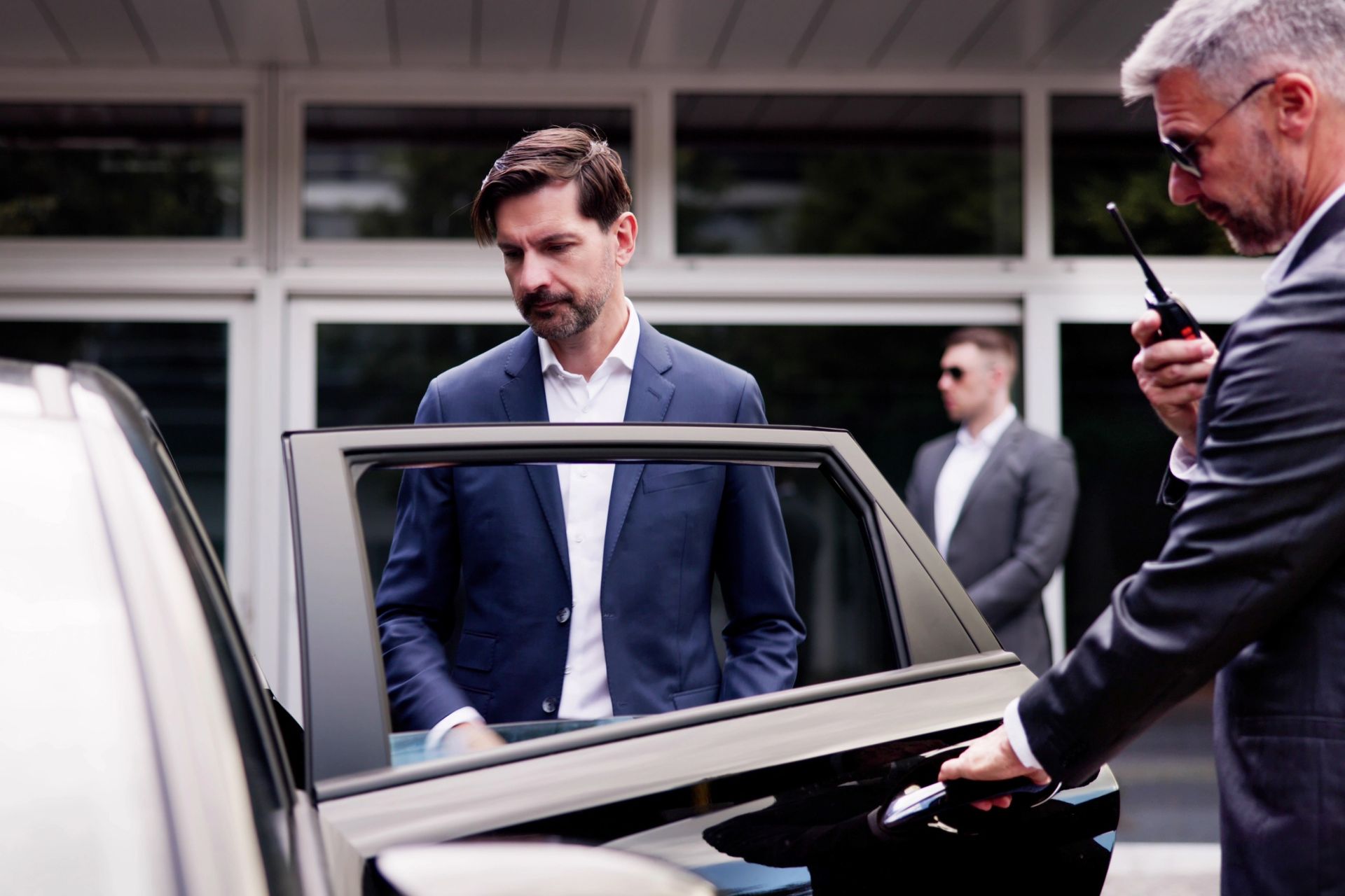 Man exiting a car, assisted by a bodyguard with a walkie-talkie; another bodyguard stands watch.