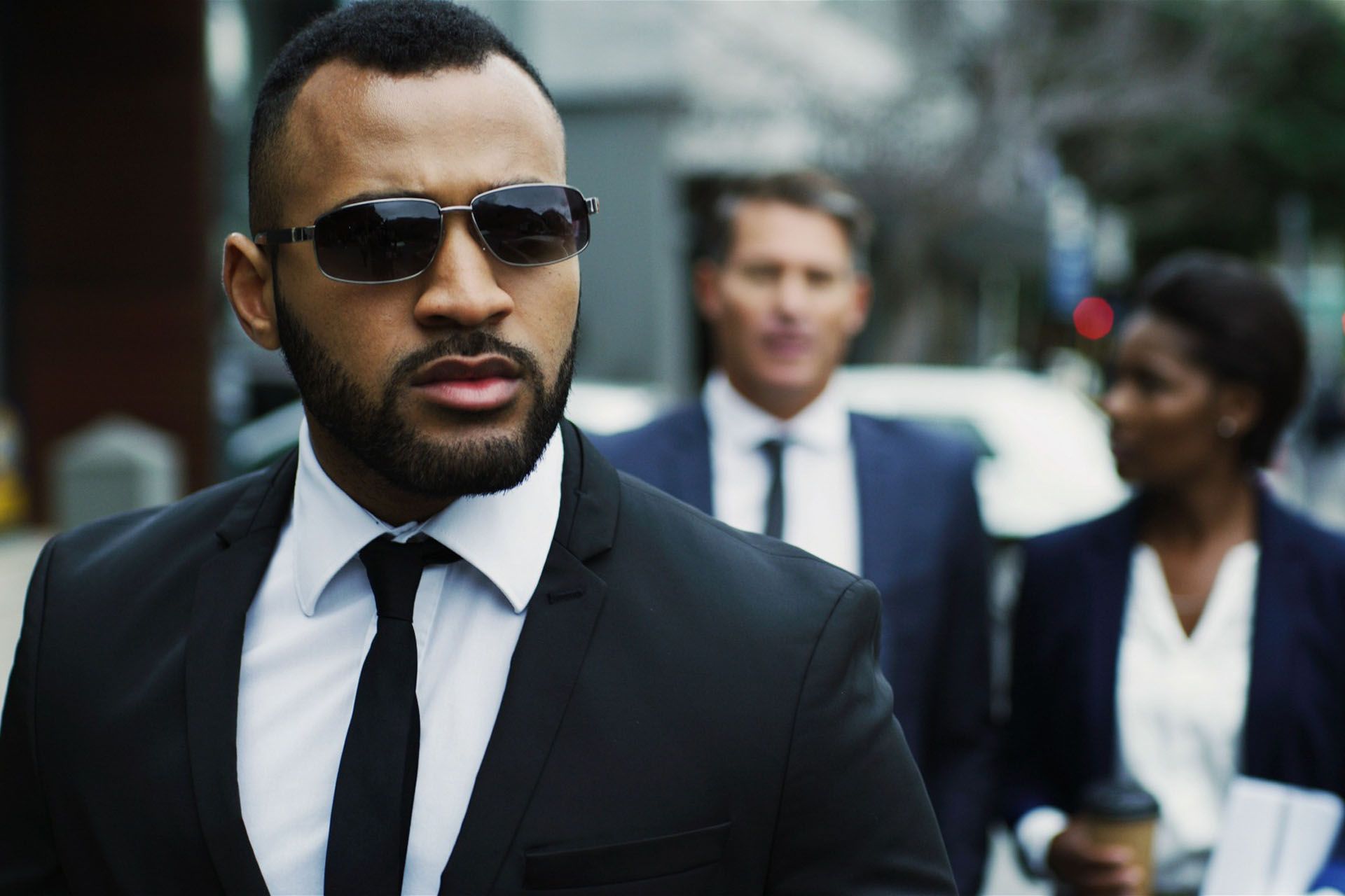 Man in black suit and sunglasses, leading two people walking on a city street.