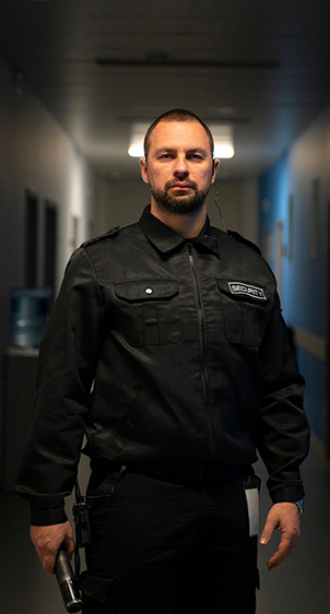 Man in black uniform, standing in a hallway, holding a gun.