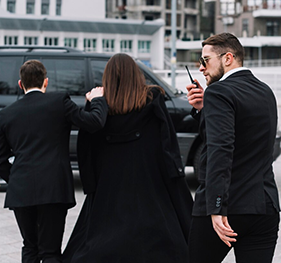 Woman escorted by two bodyguards toward a black car, one holding a walkie-talkie.