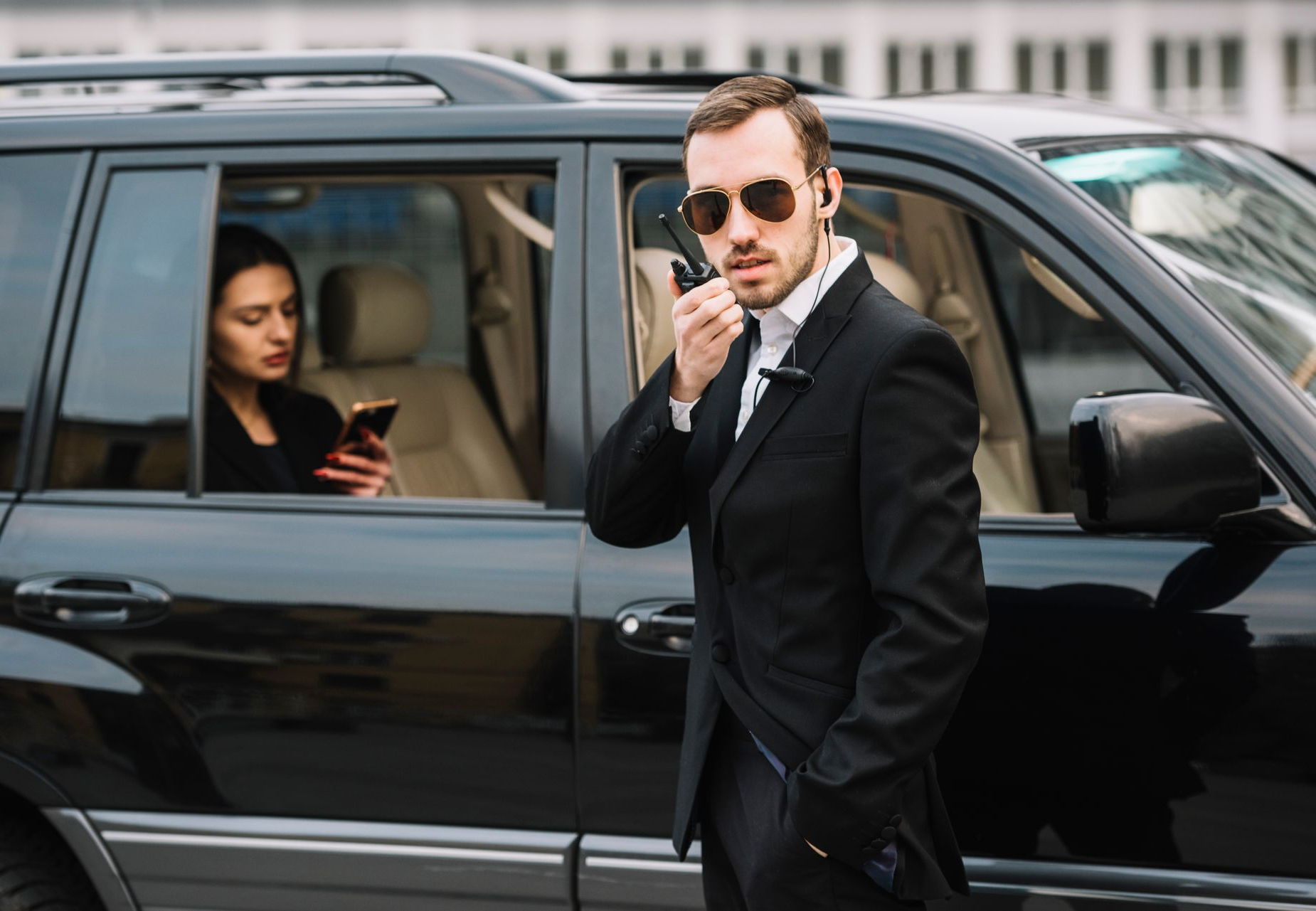 Bodyguard in black suit talking on a radio, standing beside a black SUV with a person in the back seat using a phone.