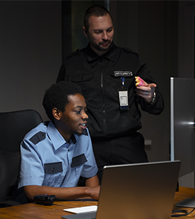 Two security guards at a desk, one looking at a laptop, the other pointing at something in his hand.