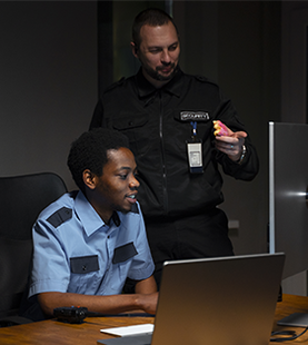 Two security guards at a desk, one looking at a laptop, the other holding something pink. Dark office setting.