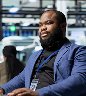 Man in a blue blazer with a lanyard, looking off-screen in a control room setting.