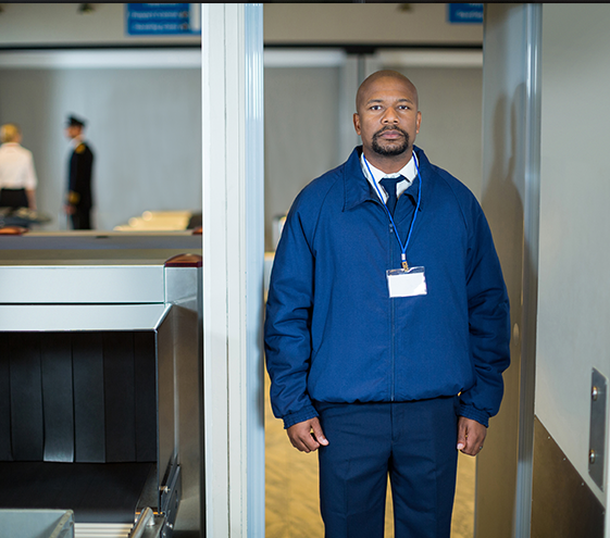 Security guard at airport checkpoint. Wearing a blue jacket and a lanyard, standing near the metal detector.