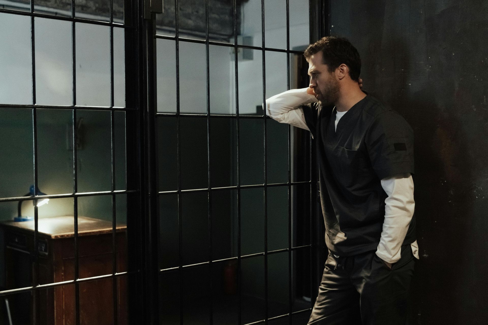 Man in scrubs leans against a prison cell door, looking dejected. Dark, industrial setting.