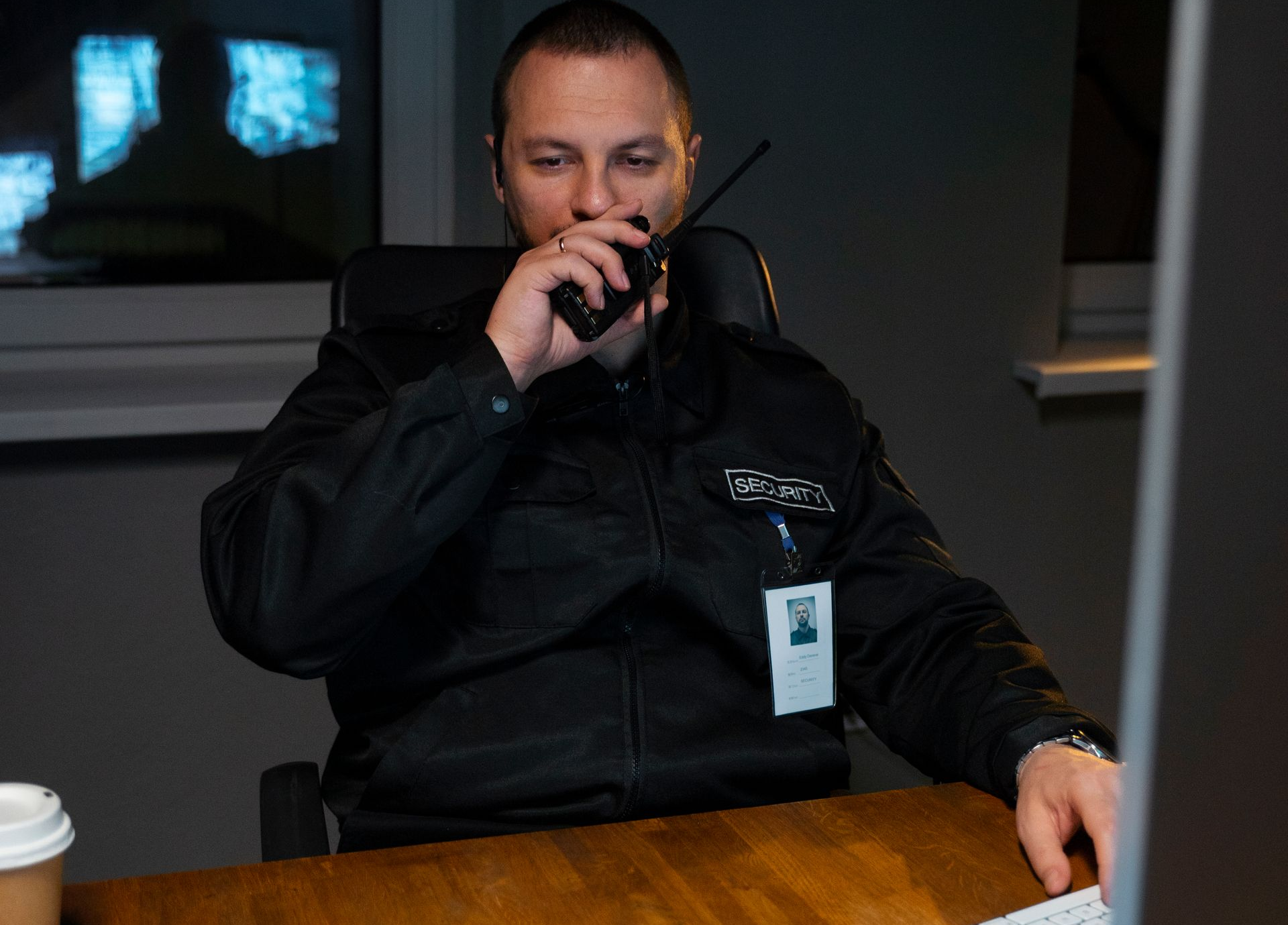 Man in a security uniform using a walkie-talkie at a desk; a cup and monitor are visible.