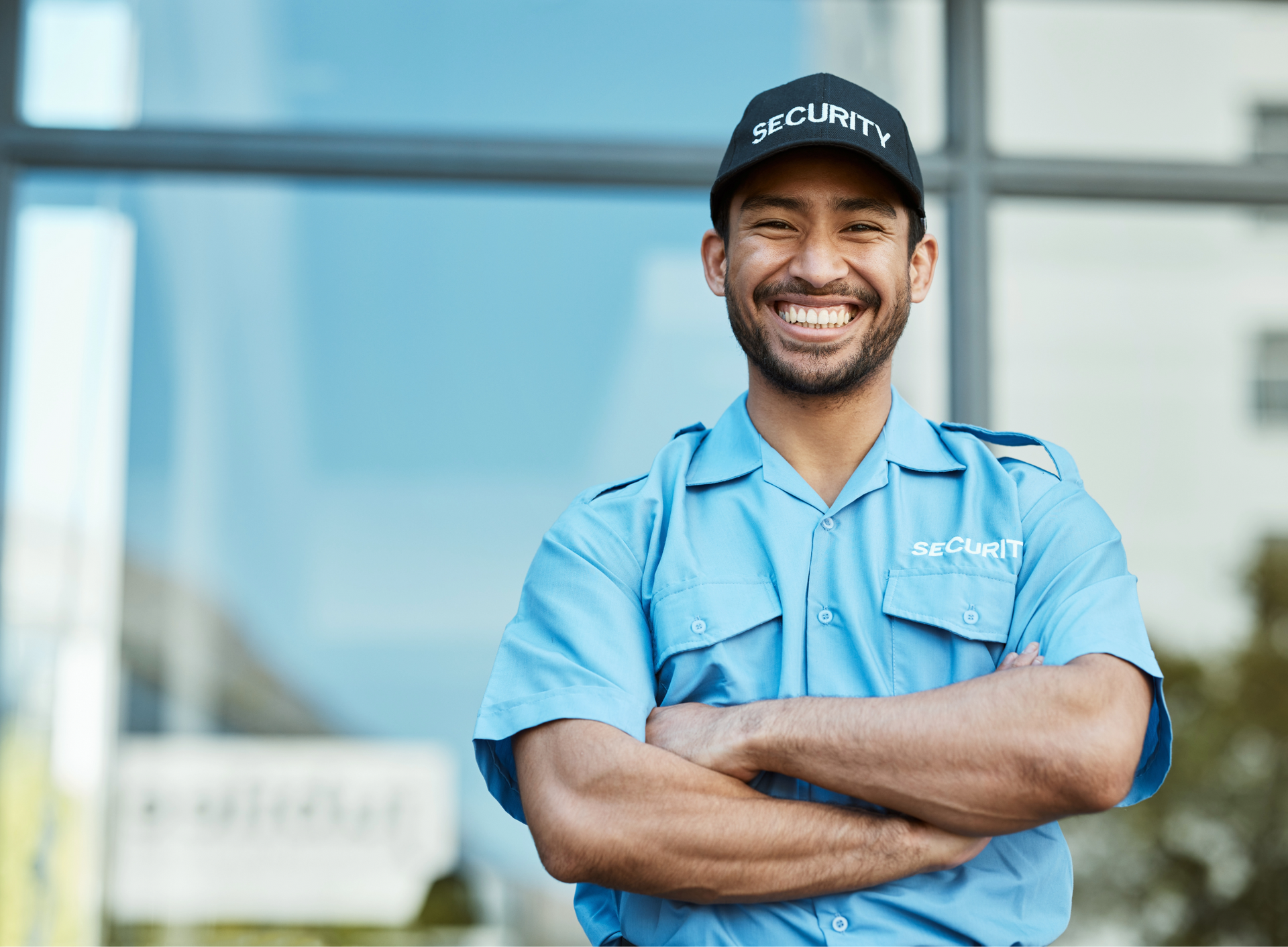 Security guard in blue uniform and cap, smiling with arms crossed in front of a building.