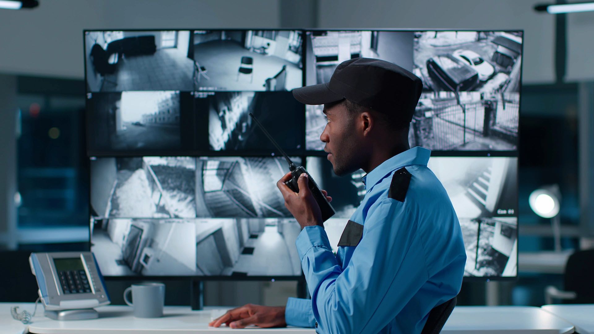 Security guard in blue uniform speaks into a walkie-talkie, monitoring security camera screens in a control room.