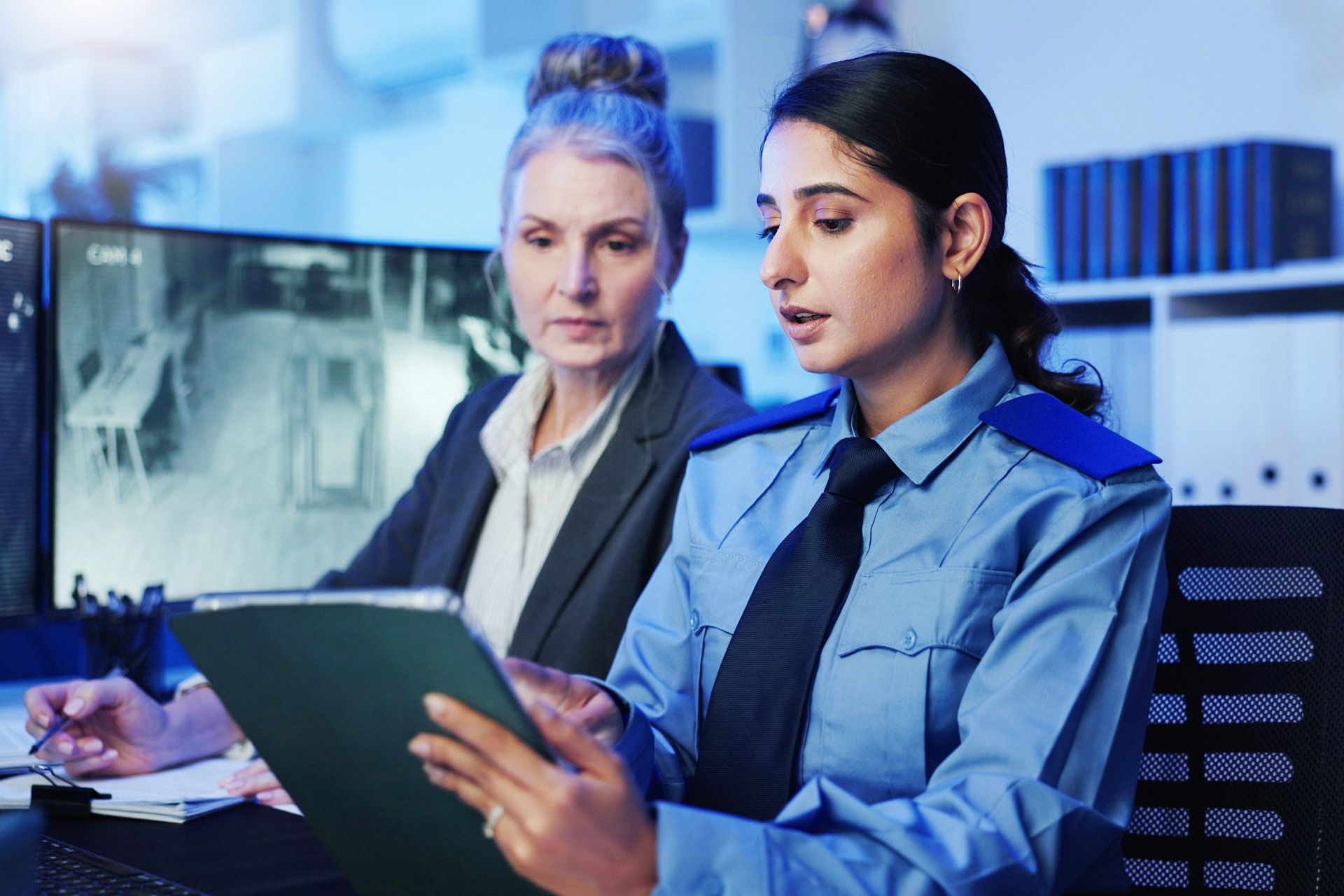 Woman in uniform shows document to another woman seated in front of security monitors.