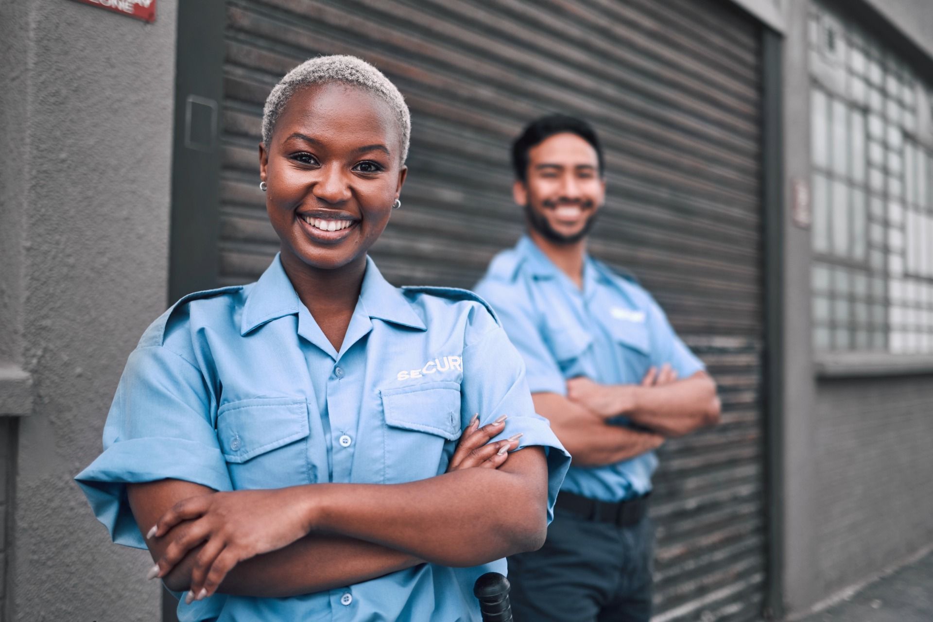 Two security guards in light blue uniforms smile, arms crossed, outside a building with a metal door.