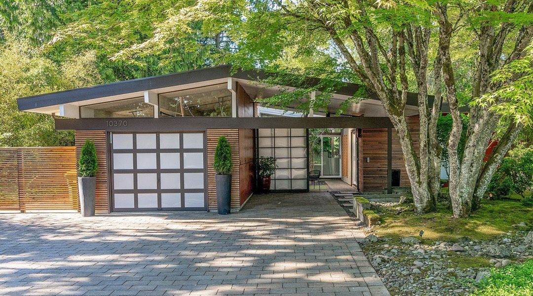 Mid-Century modern home with a dark roof and garage, light wood siding, glass entry, and a tree on the right.