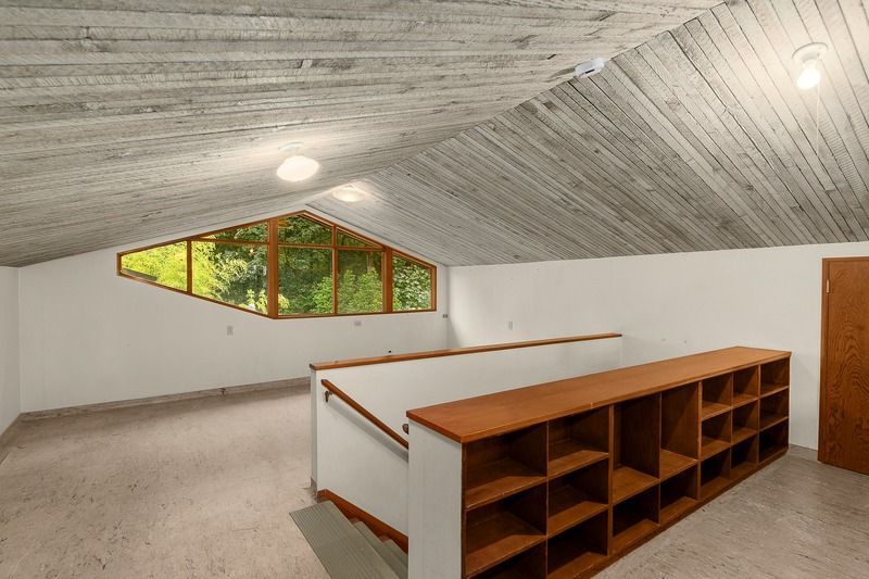 Interior view of a light-filled loft space with angled ceiling, long shelves, and a geometric window with a forest view.