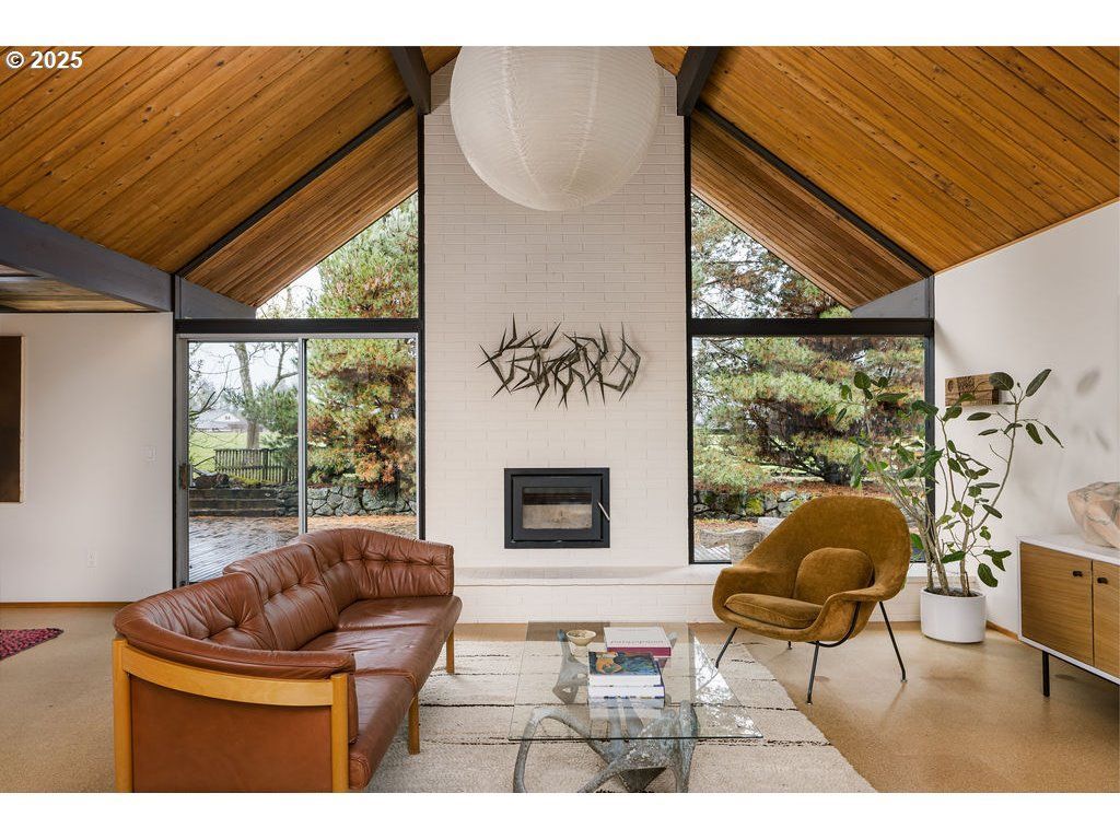 Mid-century modern living room with a brown leather sofa, fireplace, and large windows with wooded views.