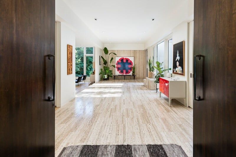 Open doorway reveals modern hallway with travertine floors, art, plants, and natural light.