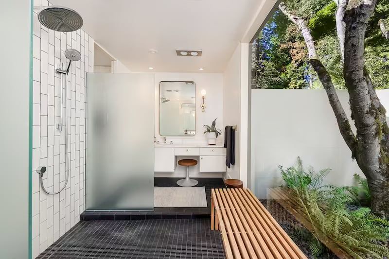 Modern bathroom with an outdoor shower. Black tiled floor and frosted glass shower divider. Bench next to green foliage.