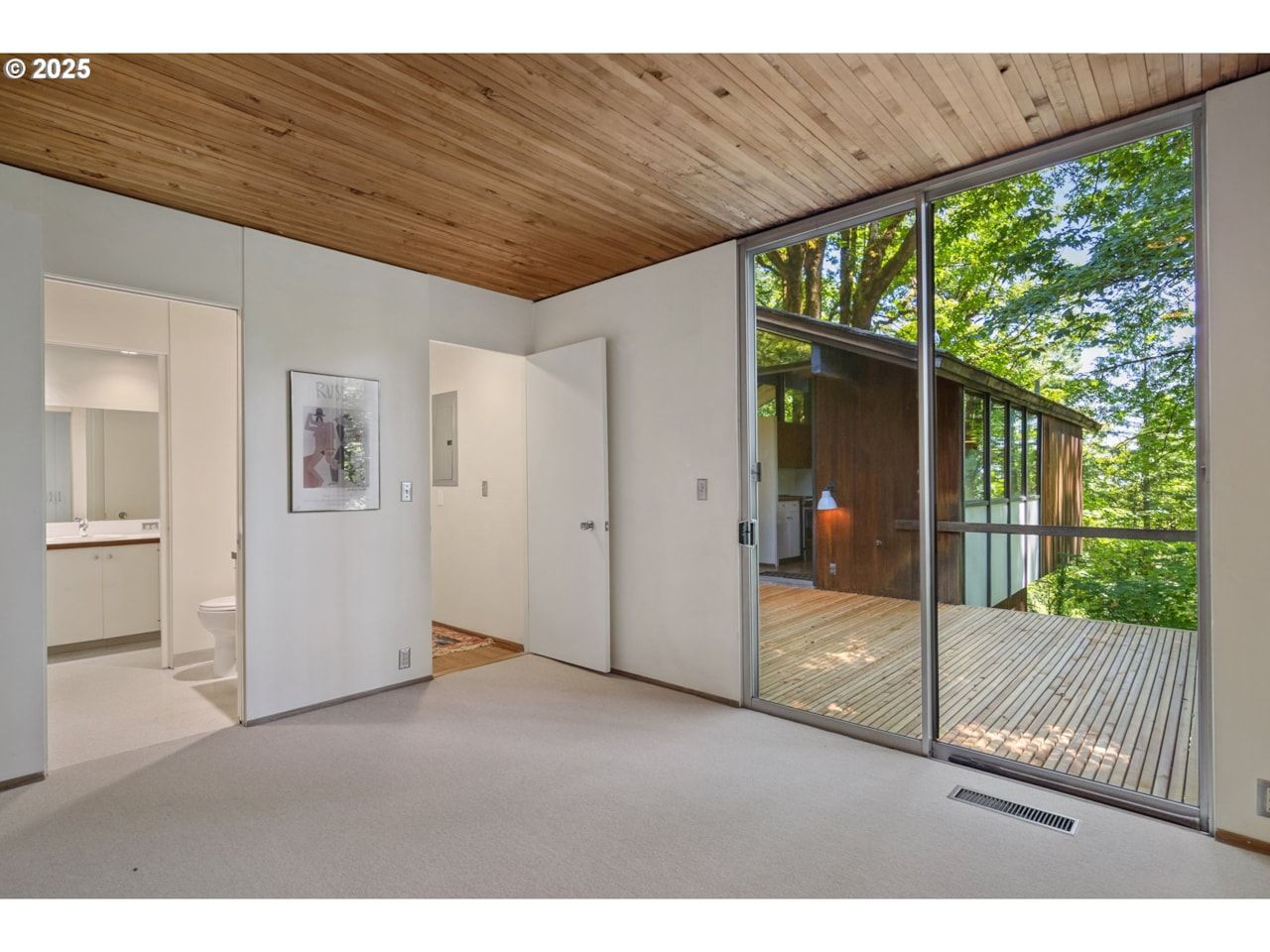 Bedroom with sliding glass doors to a deck, white walls, wood ceiling, and a view of trees.