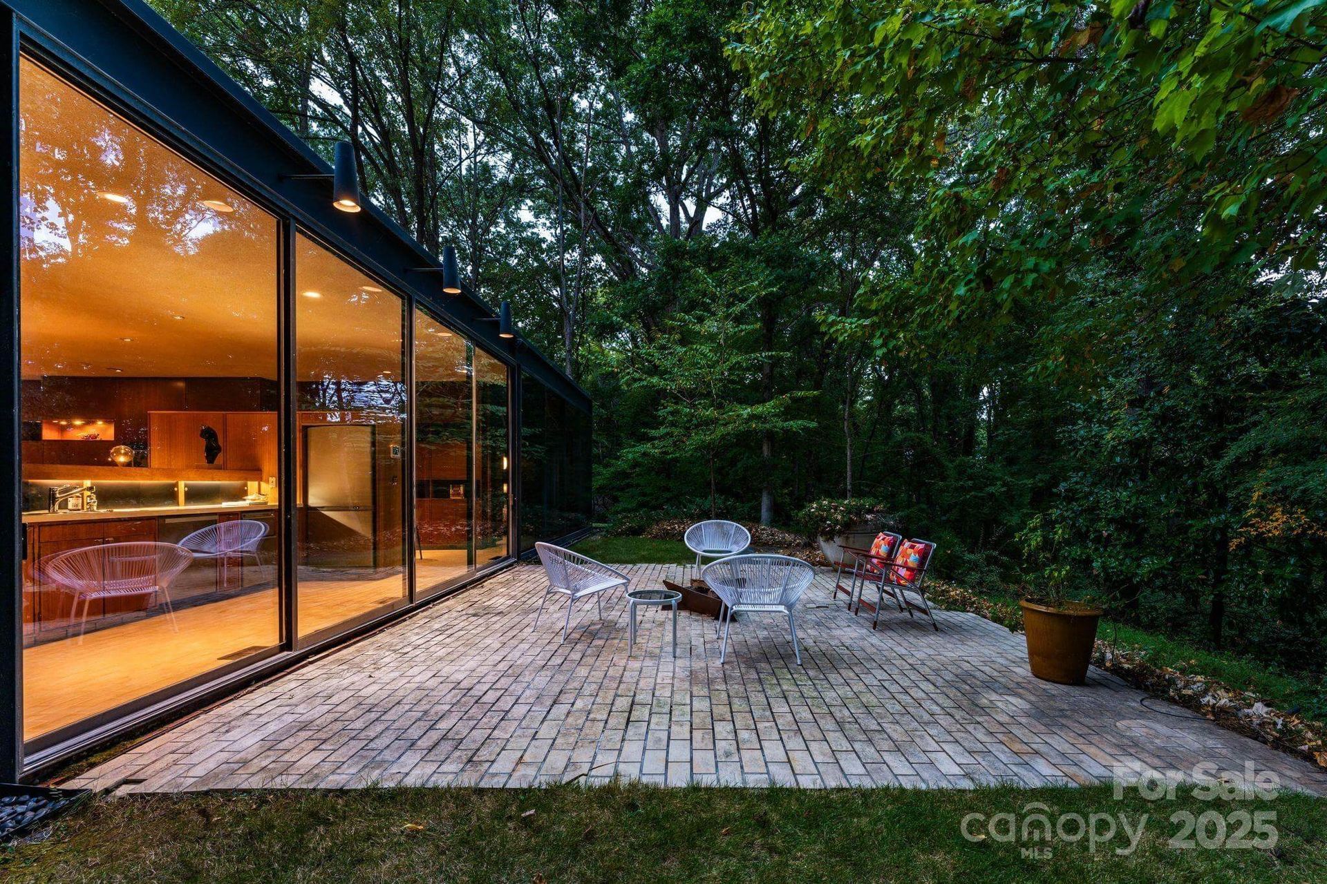 Modern house with glass walls overlooks a brick patio with seating; surrounded by trees.