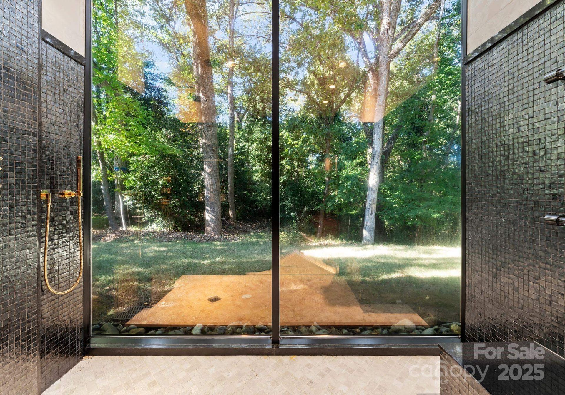 Shower with floor-to-ceiling window overlooking a lush green yard and trees; black tiled walls.