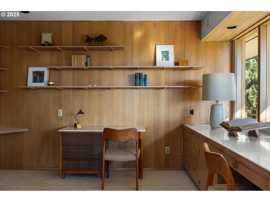 Wood-paneled home office with desk, built-in shelves displaying art and decor, and large window.