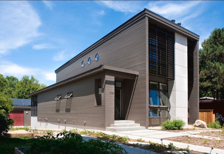 Modern two-story house with dark brown siding, large windows, and a flat roof against a blue sky.