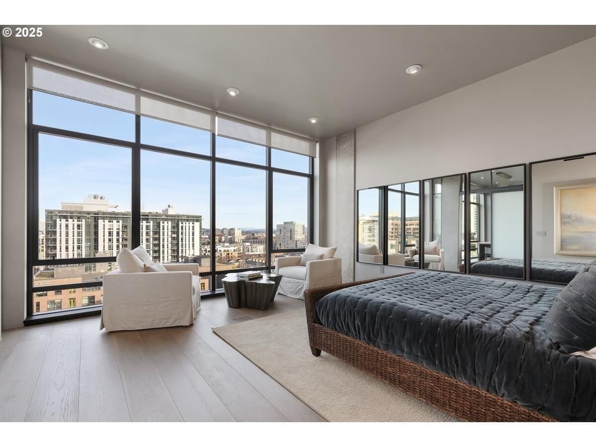 Spacious condo bedroom with large windows, city view, bed, armchairs, mirrors, neutral tones.