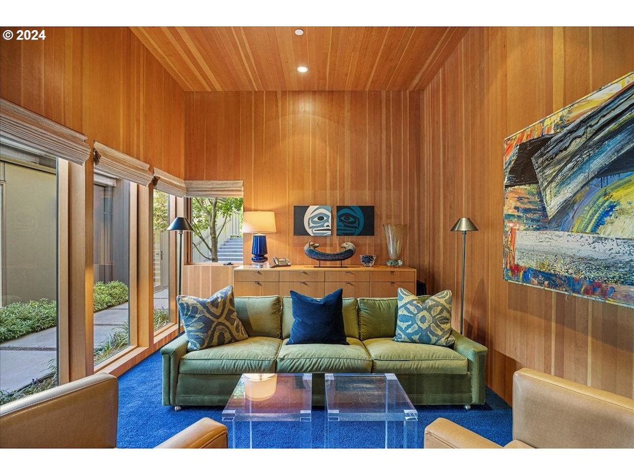Living room with wood paneling, green sofa, blue rug, acrylic coffee table, and large windows.